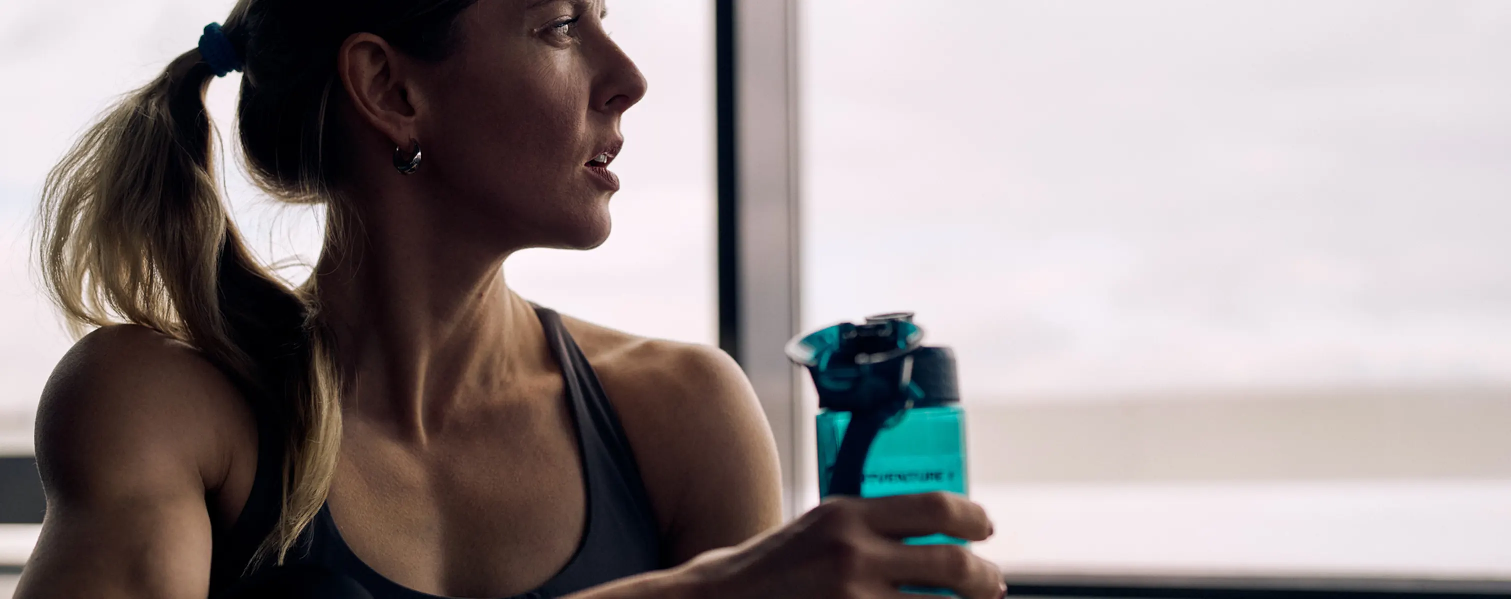 A women in workout clothes staring out a window with a water bottle in hand.