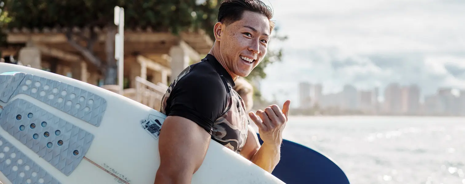 A man smiling and giving a Shaka sign with his hand, while holding a surfboard by water.