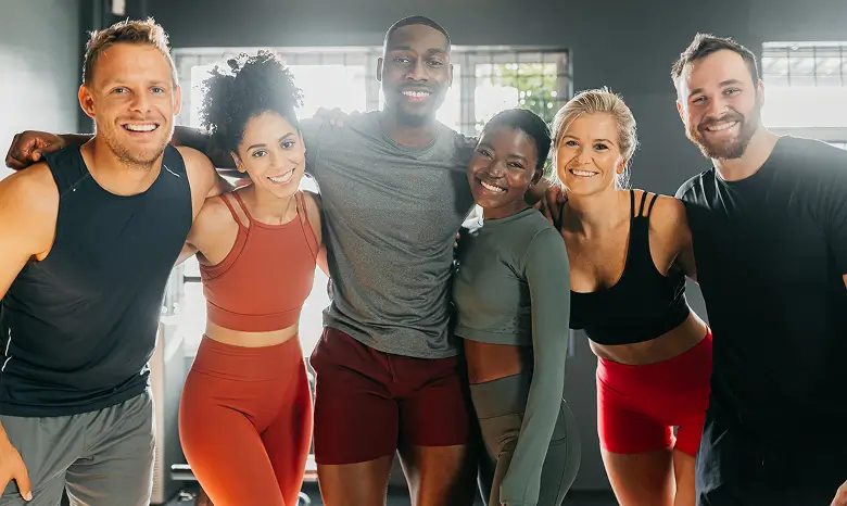 A group of smiling men and women in exercise clothes in a gym, taking a self-portrait photograph. Mobile