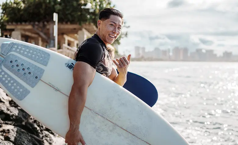 A man smiling and giving a Shaka sign with his hand, while holding a surfboard by water. Mobile